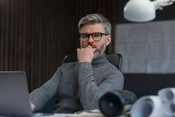 Designer working in office with laptop. Architect thinks over architectural plan, searching new ideas for construction project. Portrait of handsome bearded man sitting at workplace. Business concept - Foto, Imagem