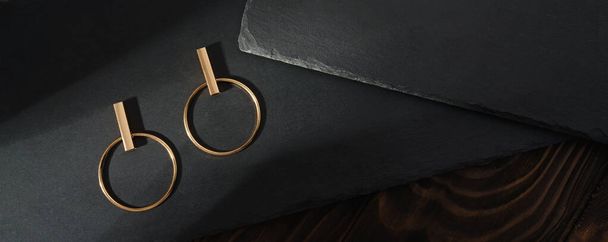 Draufsicht auf Design goldene Ohrringe auf schwarzen Steinplatten auf Holzoberfläche - Foto, Bild