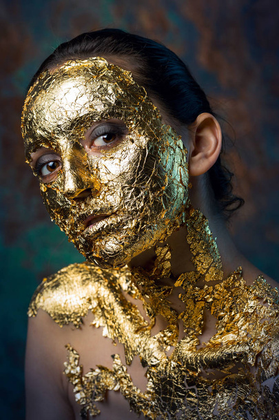 Girl with a mask on her face made of gold leaf. Gloomy studio portrait of a brunette on an abstract background. - Photo, image