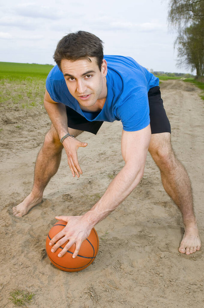 frontal wide-angle shot of a young man in a blue t-shirt,short black sports pants and barefoot bent in crouched posture,one hand resting on his knee,his other hand looking at the camera on the basketball in front of him - Foto, Imagem
