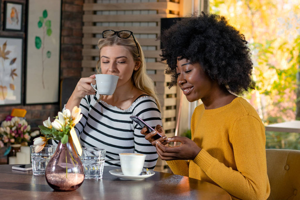 Two multi ethnic girl friends enjoying coffee together in a coffee shop, sitting at a table and looking pictures or social media content on a smartphone. - Photo, Image