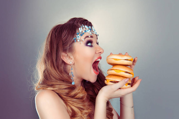 Gyorséttermi szerelem. Egy gyönyörű, vicces fiatal lány szürke lila háttérrel, szőke, tálcás hamburgerrel a kezében, három hamburgert fog harapni.. - Fotó, kép