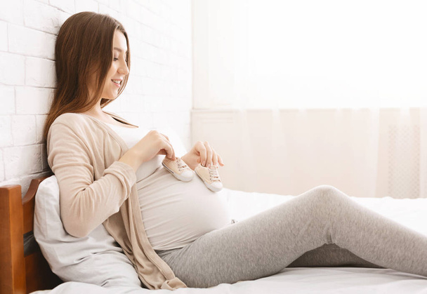 Ευτυχισμένη έγκυος γυναίκα παίζει με την κοιλιά και τις μικροσκοπικές μπότες της - Φωτογραφία, εικόνα
