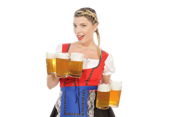 young attractive woman with blond hair and pigtails wearing a colorfull dirdl dress and holds several full beer mugs with cold beer in hand,isolated against white background. - Photo, image