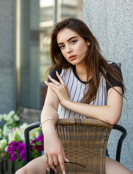 Elég szexi fiatal hipszter nő egy pólóban i egy szalmaszékben a szabadban egy kávézóban egy nyári napon. barna lány modell. - Fotó, kép