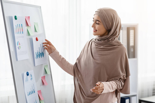Счастливая арабская предпринимательница смотрит на графики и диаграммы в офисном совете
 - Фото, изображение