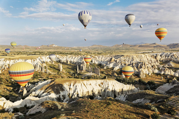 Aerial landscape of earlymorning sun falling of the rough Cappadocia terain whilst overhead the blue skies and clouds are filled with mutli colored hot air balloons - 写真・画像