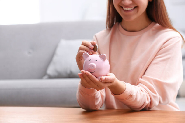 Woman putting coin into piggy bank at wooden table, closeup - 写真・画像