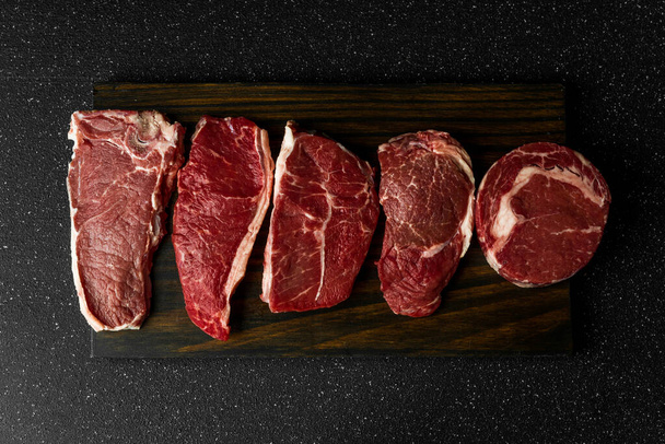 Selection of raw beef meat food steaks against black stone background. - Фото, изображение