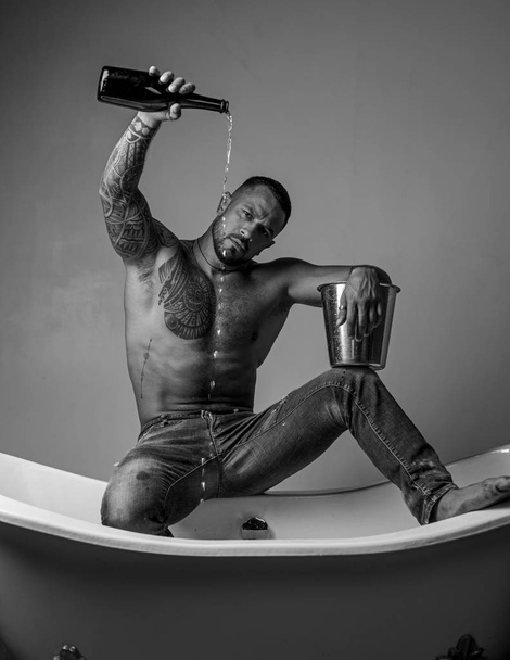 Δυνατός μυώδης άντρας με τατουάζ που κρατάει μπουκάλι σαμπάνιας και ποζάρει στο μπάνιο. Όμορφος γενειοφόρος άντρας χωρίς πουκάμισο σε τζιν με σέξι σώμα στο μπάνιο. Σεξουαλικός άντρας στο μπάνιο.. - Φωτογραφία, εικόνα