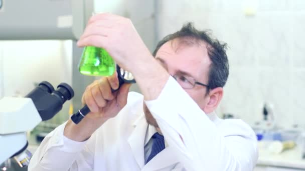 Chemist crazy. A mad scientist conducts experiments in a scientific laboratory. With surprised eyes, he looks through a magnifying glass at the green liquid in the flask. - Video