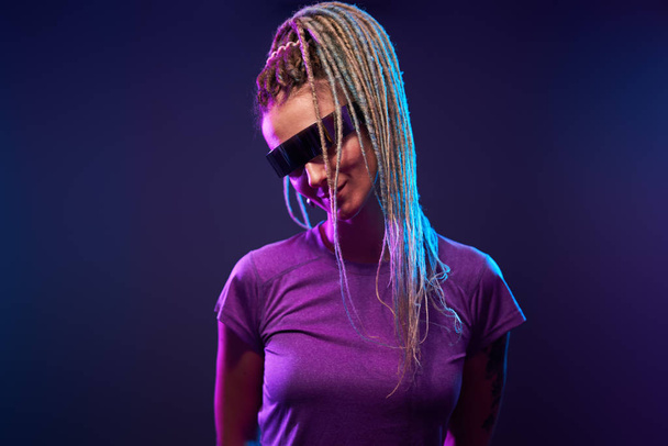 Nainen, jolla on saparot aurinkolaseissa, katselee kameraa neonvalossa
 - Valokuva, kuva
