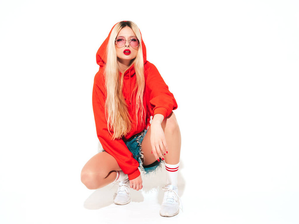 Portrait of young beautiful hipster bad girl in trendy red summer hoodie and earring in her nose.Sexy carefree smiling blond woman sitting in studio.Positive model having fun.Isolated on white - 写真・画像