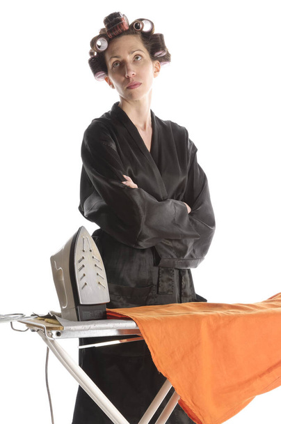adult housewife with curlers in her hair and a bathrobe stands behind the ironing board and makes with the iron ironing in front of a white background. - Fotoğraf, Görsel
