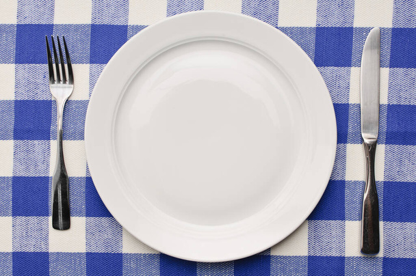 white pozellanteller on blue-white checked tablecloth with knife and fork. - Foto, Imagem