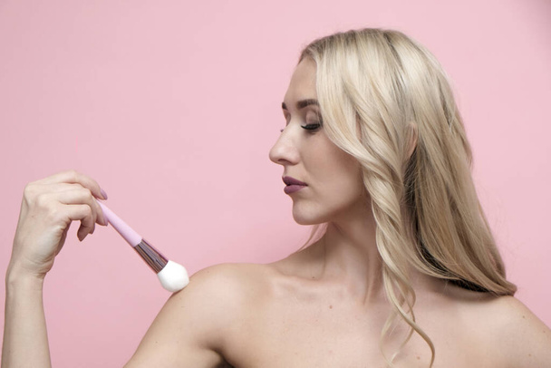 Beautiful blond woman with naked shoulders holding brush on pastel pink background. Beauty, cosmetics and spa concept - Photo, Image