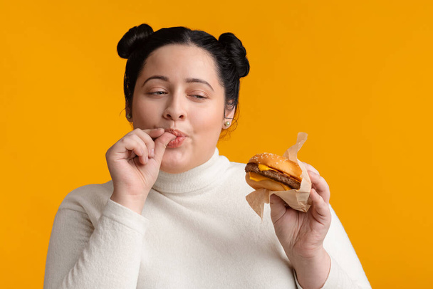 Συν το μέγεθος κορίτσι τρώει σάντουιτς τσίζμπεργκερ και γλείφει δάχτυλα με όρεξη - Φωτογραφία, εικόνα