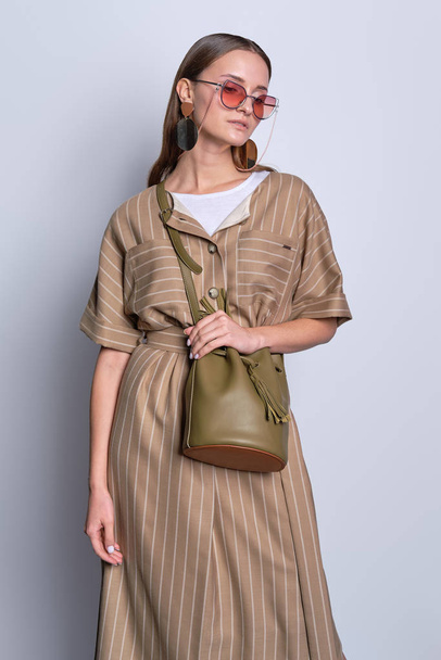Trendy lady in big sunglasses wearing striped olive dress with leather handbag posing over gray background - Foto, afbeelding