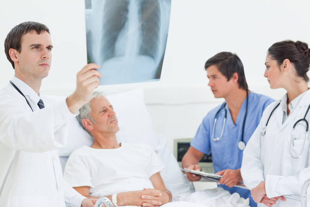 Doctor looking at an x-ray scan of lungs while standing next to the bed of a patient who is looking at a doctor and a nurse - Photo, Image