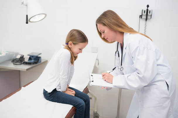 Doctor writing in a notepad in front of a smiling girl sitting in an examination room - Photo, Image