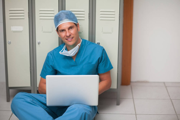Smiling surgeon sitting on the floor of a locker while holding a laptop in a hospital - Fotoğraf, Görsel