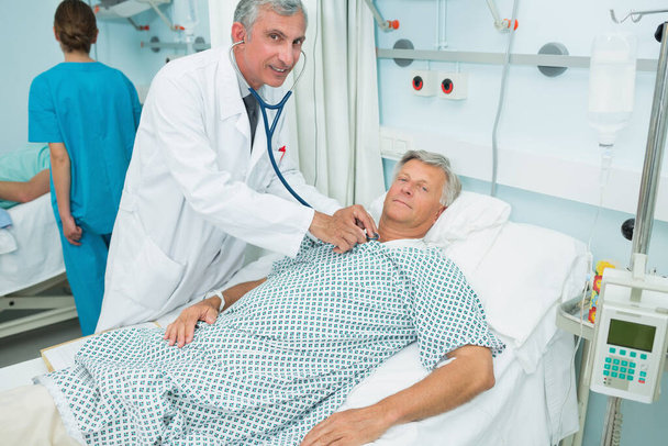 Smiling male doctor auscultating the chest of a male patient in a bed ward in a hospital - Photo, Image
