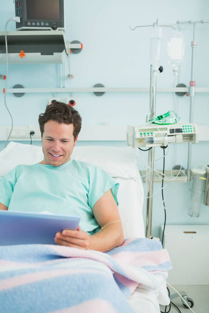 Smiling male patient using a tactile tablet while lying on a bed in a hospital - Valokuva, kuva