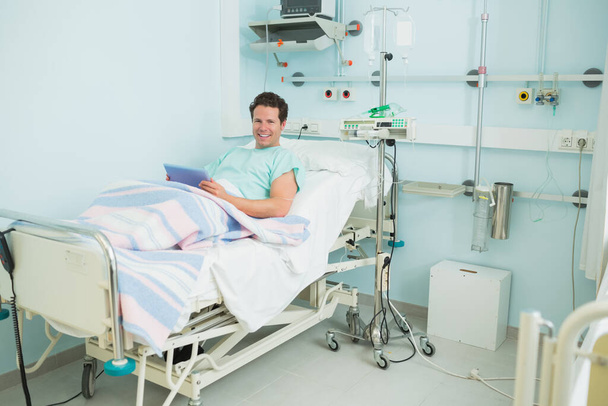 Male patient smiling while holding a tactile tablet while lying on a bed in a hospital ward - Valokuva, kuva