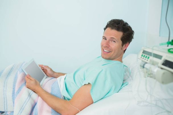Усміхнений пацієнт чоловічої статі торкається тактильної таблетки, лежачи на ліжку в лікарняному палаті
 - Фото, зображення