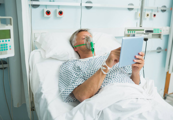 Patient lying on a bed with a mask while holding a tactile tablet in a hospital room - Fotó, kép