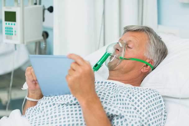 Senior male patient lying on a bed with a mask while holding a tactile tablet in a hospital room - Foto, imagen