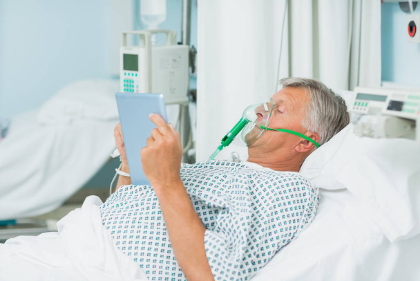 Male patient wearing an oxygen mask while holding a tactile tablet in a hospital bed - 写真・画像