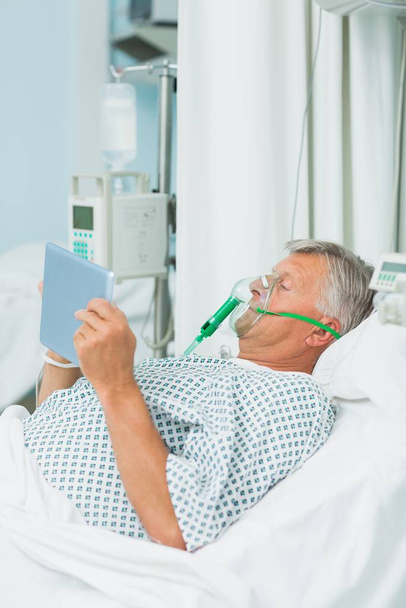 Senior male patient wearing an oxygen mask while using a tactile tablet in a hospital bed - Zdjęcie, obraz