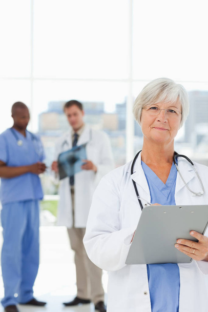 Mature doctor with clipboard and talking colleagues behind her - Fotoğraf, Görsel
