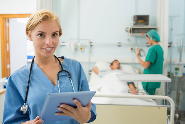 Blonde nurse holding a tablet computer while smiling in hospital ward - Foto, afbeelding