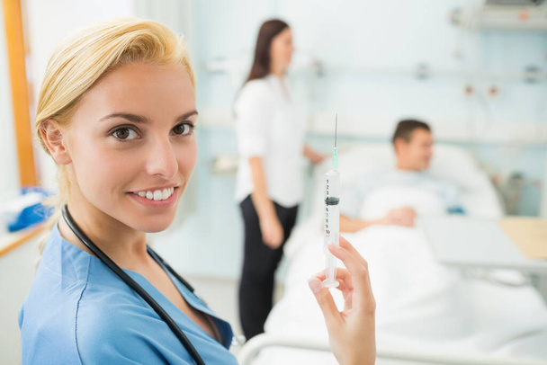 Медсестра держит шприц, улыбаясь в больничной палате - Фото, изображение