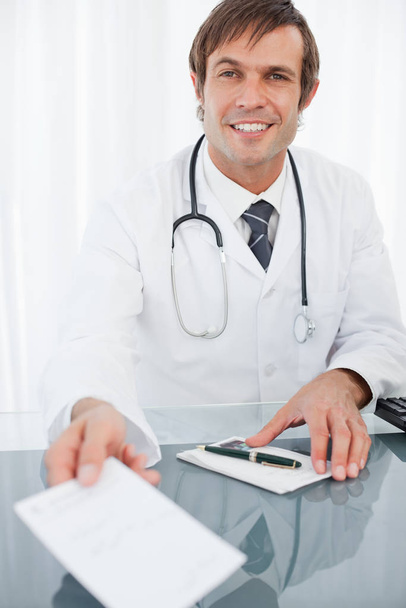 Smiling doctor holding out a prescription while sitting at his desk - Fotó, kép