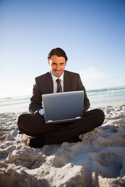 Smiling businessman working with a laptop while sitting cross-legged on the beach - Fotografie, Obrázek
