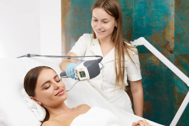 beautiful woman with smile on fcae receiving facial treatment with laser in beauty clinic, pretty girl getting face procedures with beautician - Valokuva, kuva