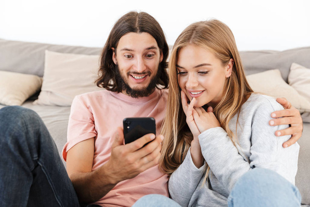 Loving couple on sofa indoors at home using mobile phone. - Φωτογραφία, εικόνα