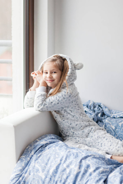 薄い灰色のパジャマの女の子が窓のそばのベッドに座って子供は朝目が覚めた - 写真・画像