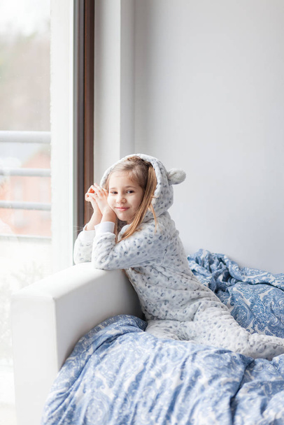 tyttö vaalean harmaa pyjama istuu sängyllä ikkunan vieressä, lapsi heräsi aamulla
 - Valokuva, kuva
