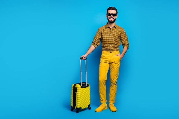 Full size photo positive bearded guy tourist enjoy air voyage stand in terminal wait check-in hold total yellow rest look trolley wear plaid clothes sneakers isolated blue color background - Photo, image