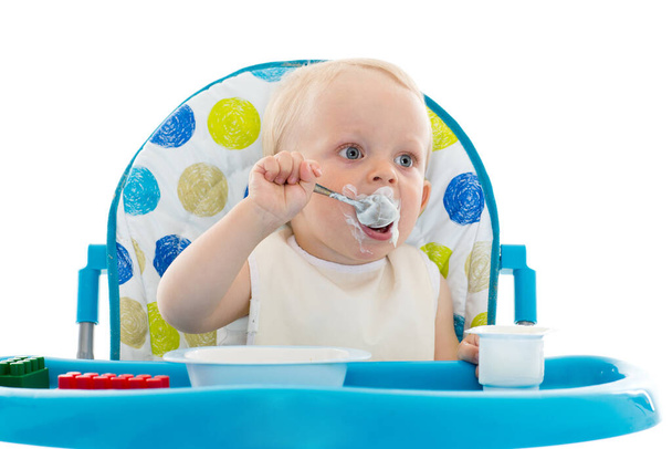 Sweet baby learning to eat with spoon sits on baby chair on a white background. - Foto, imagen