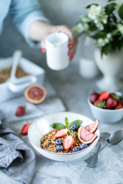 Zdrowe śniadanie z granolą, jogurtem, owocami, jagodami na białym talerzu w białym talerzu. Wybiórcze skupienie. Kobieta wlewa mleko do granoli.. - Zdjęcie, obraz