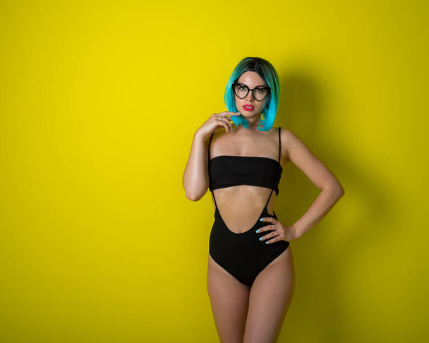 Beautiful woman in a black bikini in a colored wig and sunglasses on a yellow background. Girl posing in the studio. - Photo, Image