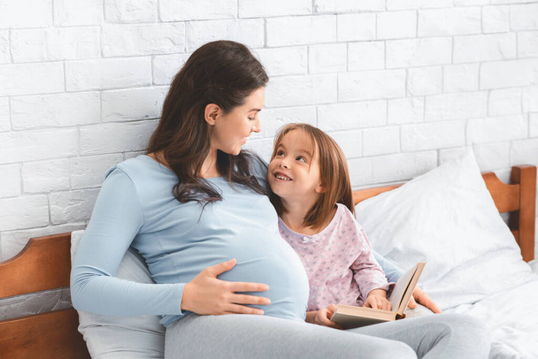 Az óvodás lány könyvet tárgyal a terhes anyjával. - Fotó, kép