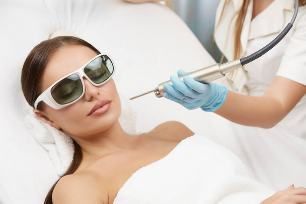 pretty femal having facial procedures and beautician holding laser near her face, beauty and cosmetology concept, hair removing by doctor in spa salon - Photo, Image