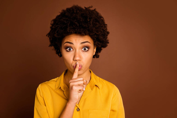 Be silent. Closeup photo of beautiful pretty dark skin lady holding finger on lips keeping secret information in privacy wear yellow shirt isolated brown color background - 写真・画像