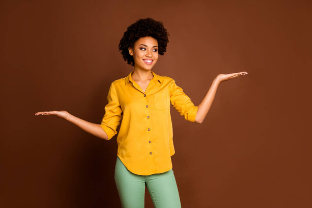 Photo of beautiful business dark skin curly lady holding open palms prefer one best variant discount novelty products wear yellow shirt green trousers isolated brown color background - Φωτογραφία, εικόνα